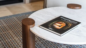 A round table with a book titled "GRANDS VINS" on a marble surface, a patterned rug, a wooden stool, a white chair, and a white cabinet with drawers. - Le Prieuré - Château La Martinette (Le Thoronet)