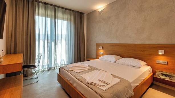 A modern hotel room with a large bed, a wooden headboard, a desk with a chair, and a large window with curtains. - Lucciola Boutique Rooms (Budva)