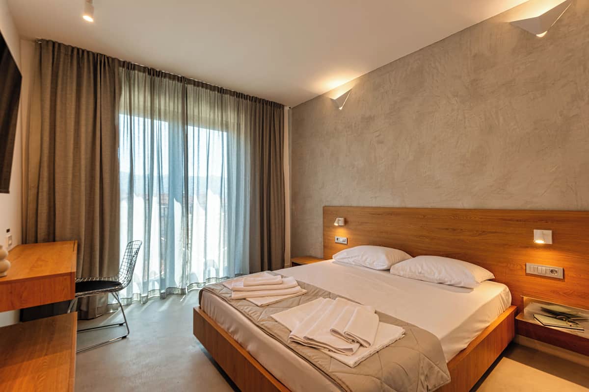 A modern hotel room with a large bed, a wooden headboard, a desk with a chair, and a large window with curtains.