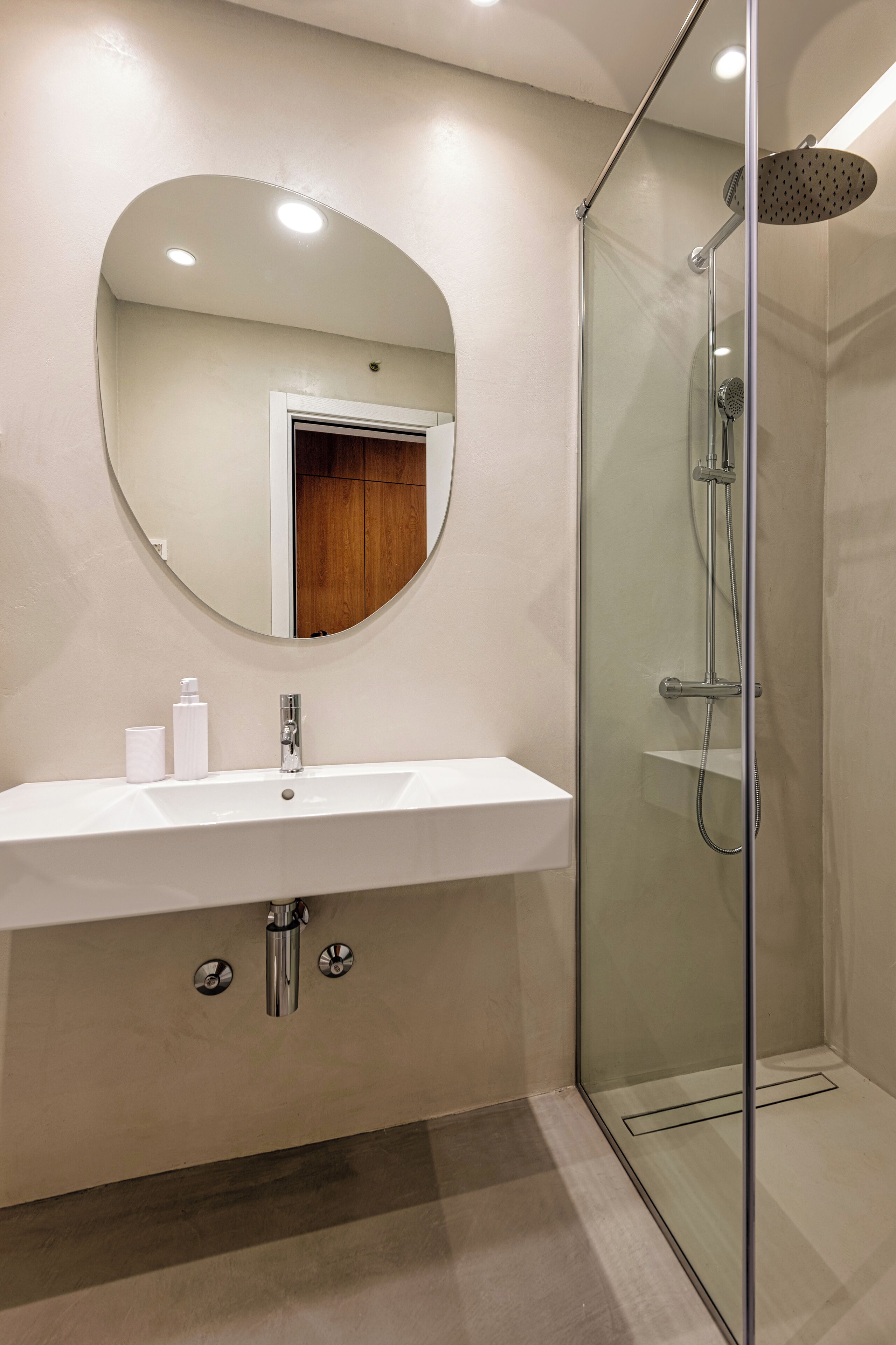 A modern bathroom with a glass-enclosed shower, a white sink with a mirror above it, and a wooden door in the background.