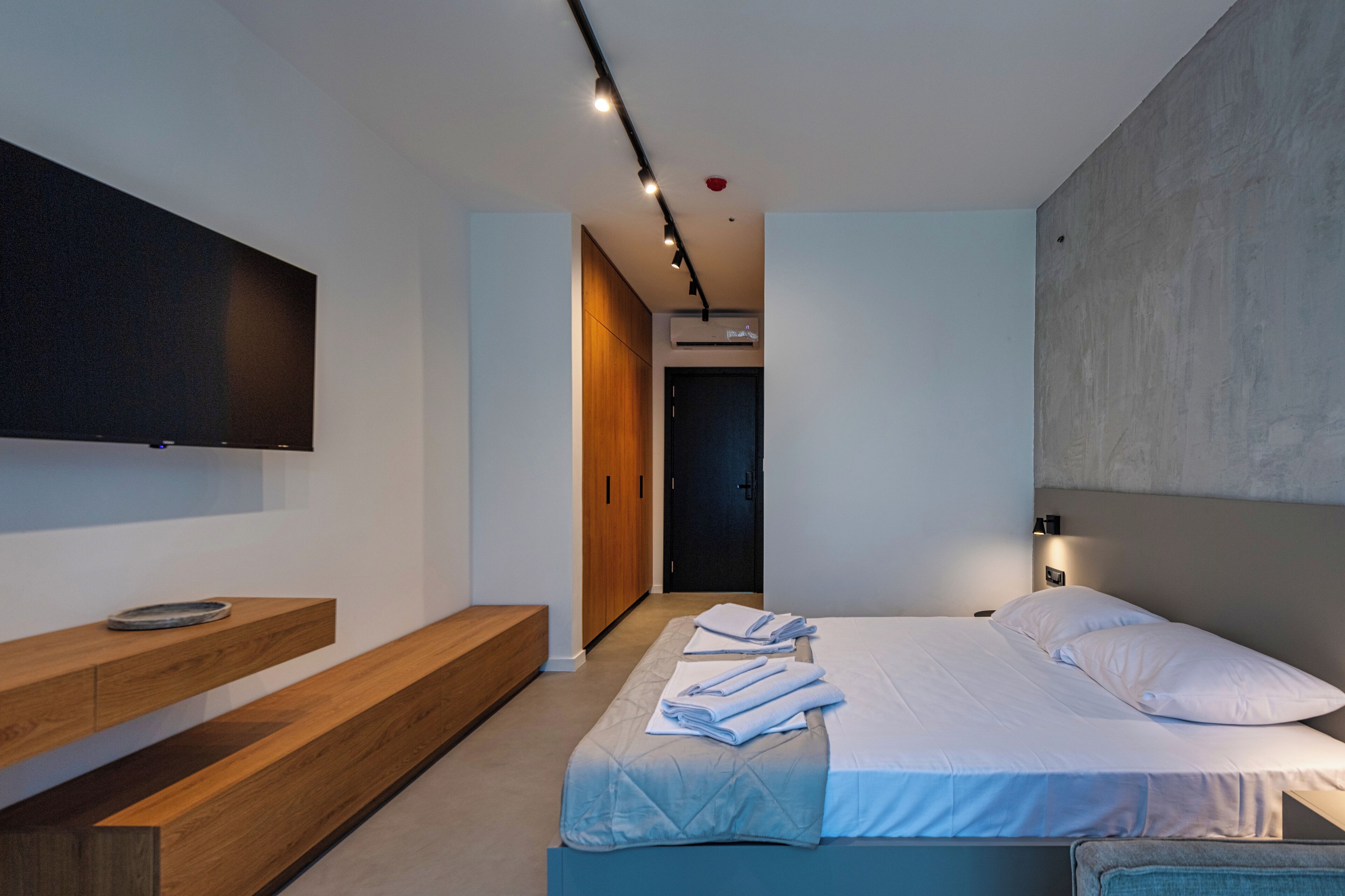 A modern hotel room with a bed, a wooden bench, a flat-screen TV, and a wall-mounted light fixture.