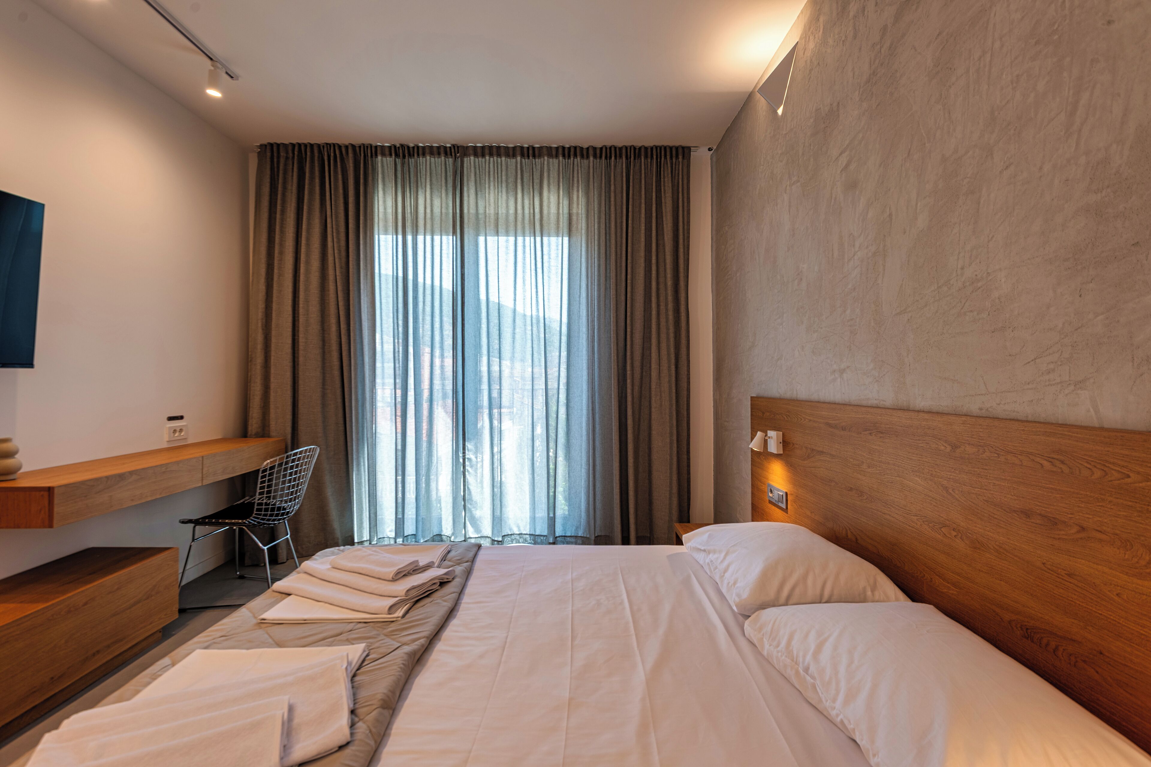 A modern hotel room with a large bed, a wooden headboard, a desk with a chair, and a large window with curtains.