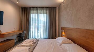 A modern hotel room with a large bed, a wooden headboard, a desk with a chair, and a large window with curtains.