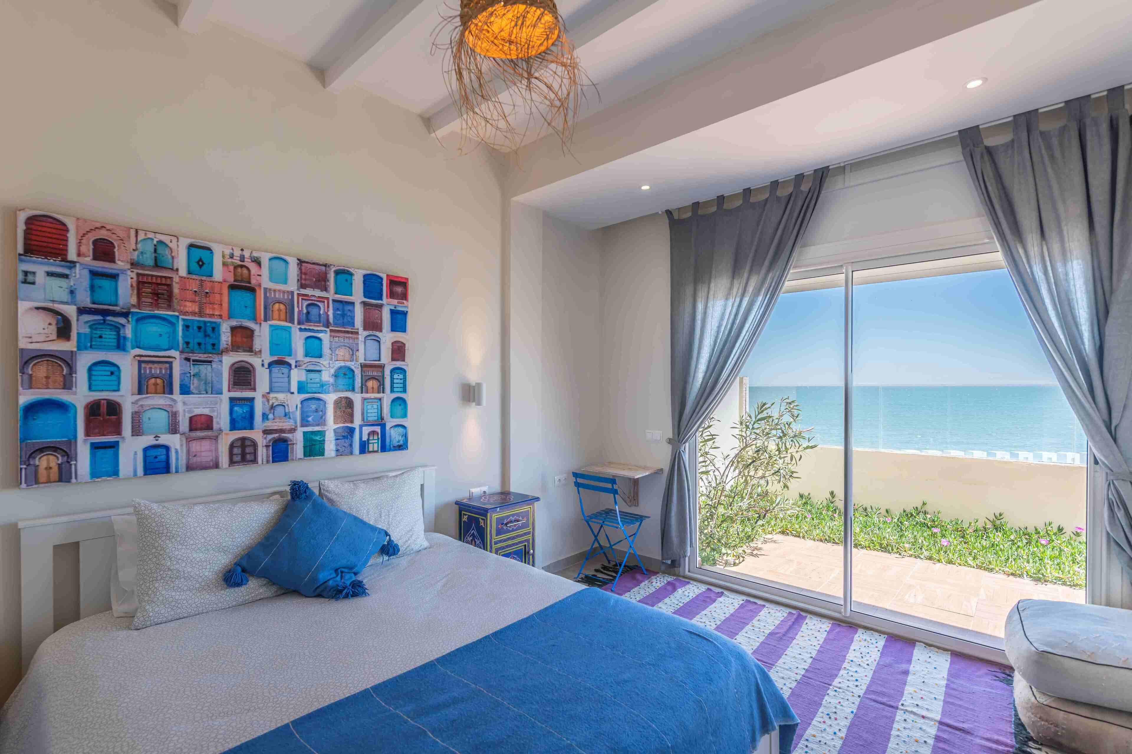 A bedroom with a large bed, a view of the beach, a wall with colorful door illustrations, and a small table with a chair.