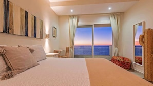 Habitación de hotel con una cama grande, un escritorio, una silla y vista al mar al atardecer.