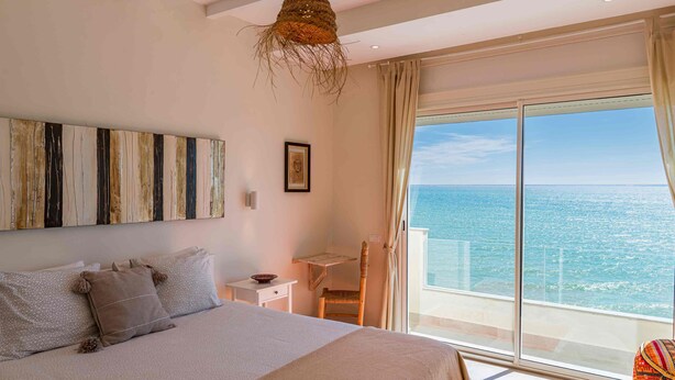 Un dormitorio con una cama grande, un escritorio pequeño, una silla y vistas al mar a través de un ventanal amplio.