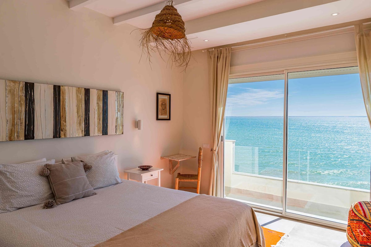 Une chambre avec un grand lit, un petit bureau, une chaise et une vue sur la mer par une grande fenêtre.