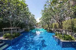 A swimming pool with tiled blue flooring, surrounded by trees and a stone wall. - The Title Rawai Pool View Apartment F303 (Rawai)
