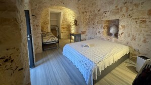 Un dormitorio con cama, una silla y una mesita. Se observa una pared de piedra y un arco que conduce a otra habitación.