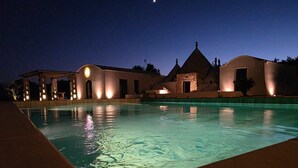 Un área de piscina iluminada por la noche, con un edificio que presenta arcos y una estructura de piedra.