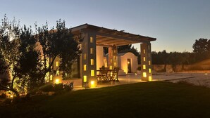 Un área de estar al aire libre techada, con una mesa y sillas, rodeada de columnas iluminadas y un muro de piedra.