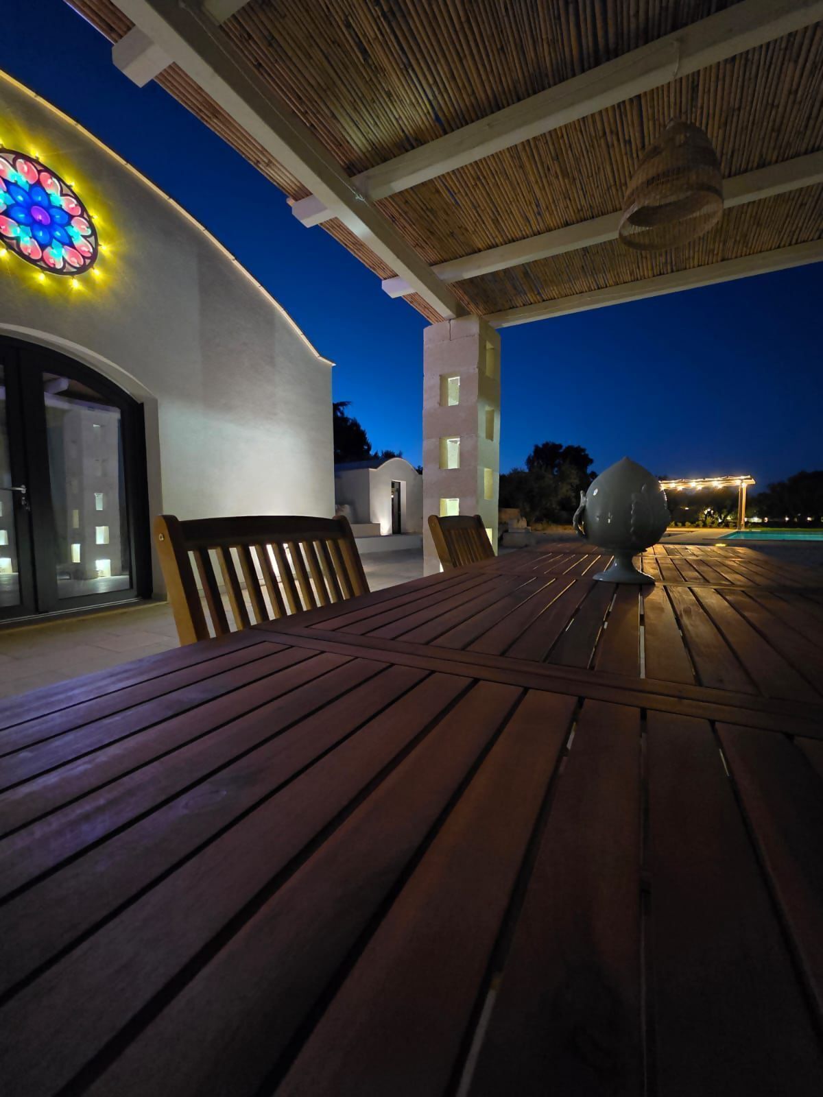 An outdoor wooden deck with a bench and a table, a building with a white facade and a stained glass window, and a clear sky at dusk.