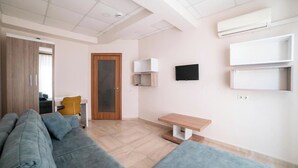 Een moderne woonkamer met een grijze bank, een klein houten tafeltje en een aan de muur bevestigde televisie.