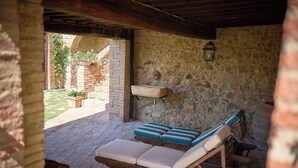 An outdoor seating area with wooden lounge chairs and cushions, a stone wall, and a hanging planter with flowers. - Agriturismo Le Case (Castiglione d'Orcia)