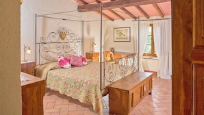 A four-poster bed with a floral bedspread and a pink pillow, a wooden desk with a chair, a wooden bench, and a window with white curtains. - Agriturismo Le Case (Castiglione d'Orcia)