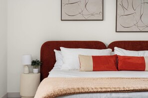 A neatly made bed with a red headboard, white pillows, and a beige throw blanket. There is a bedside table with a lamp and a small plant. - Upscale Zilker Gem | Pool & Two Kitchens (Austin)