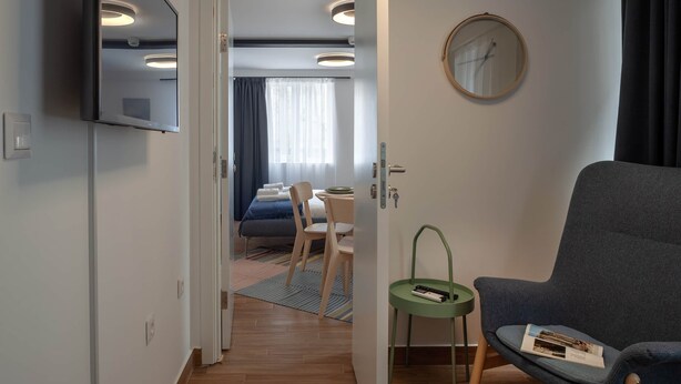 Ein modernes Hotelzimmer mit einem Flachbildfernseher, einem runden Spiegel, einer Sitzecke mit Sessel und kleinem Tisch sowie Blick in das angrenzende Schlafzimmer.