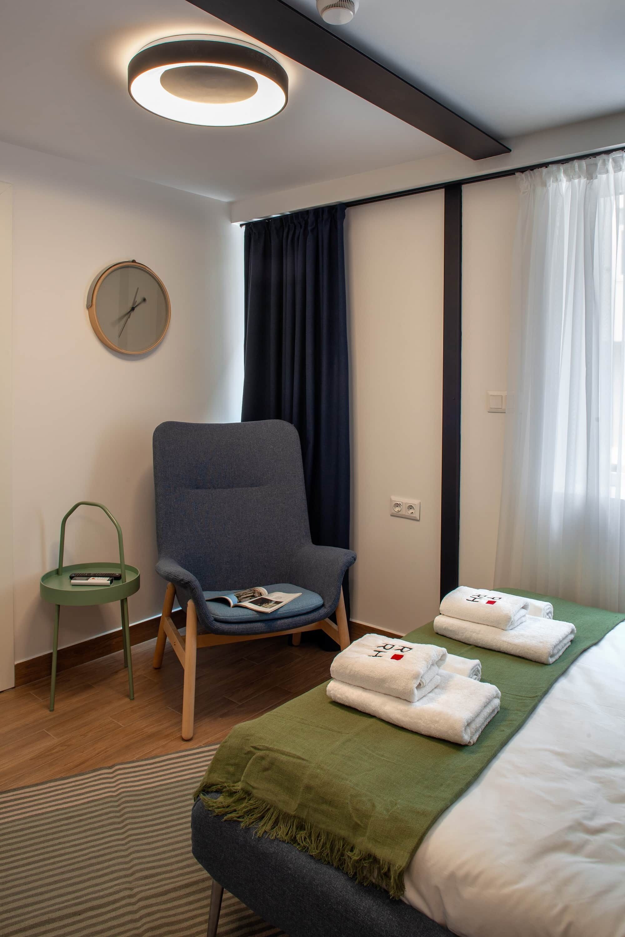 Habitación de hotel con cama, una silla, una mesita, un reloj y una ventana con cortinas.