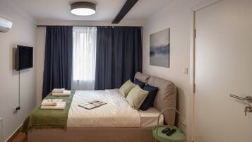 A bedroom with a bed, a bedside table with a remote, a wall-mounted TV, a window with curtains, and a door.