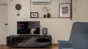 Ein modernes Wohnzimmer mit einem Flachbildfernseher, einem blauen Sessel, einem Couchtisch mit Croissants und Tassen sowie einer Wand-Klimaanlage.