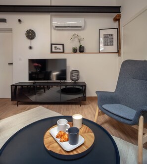 A modern living room with a flat-screen TV, a blue armchair, a coffee table with croissants and cups, and a wall-mounted air conditioner.