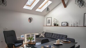 A modern living room with a dining table, grey sofas, and a large window with skylights.