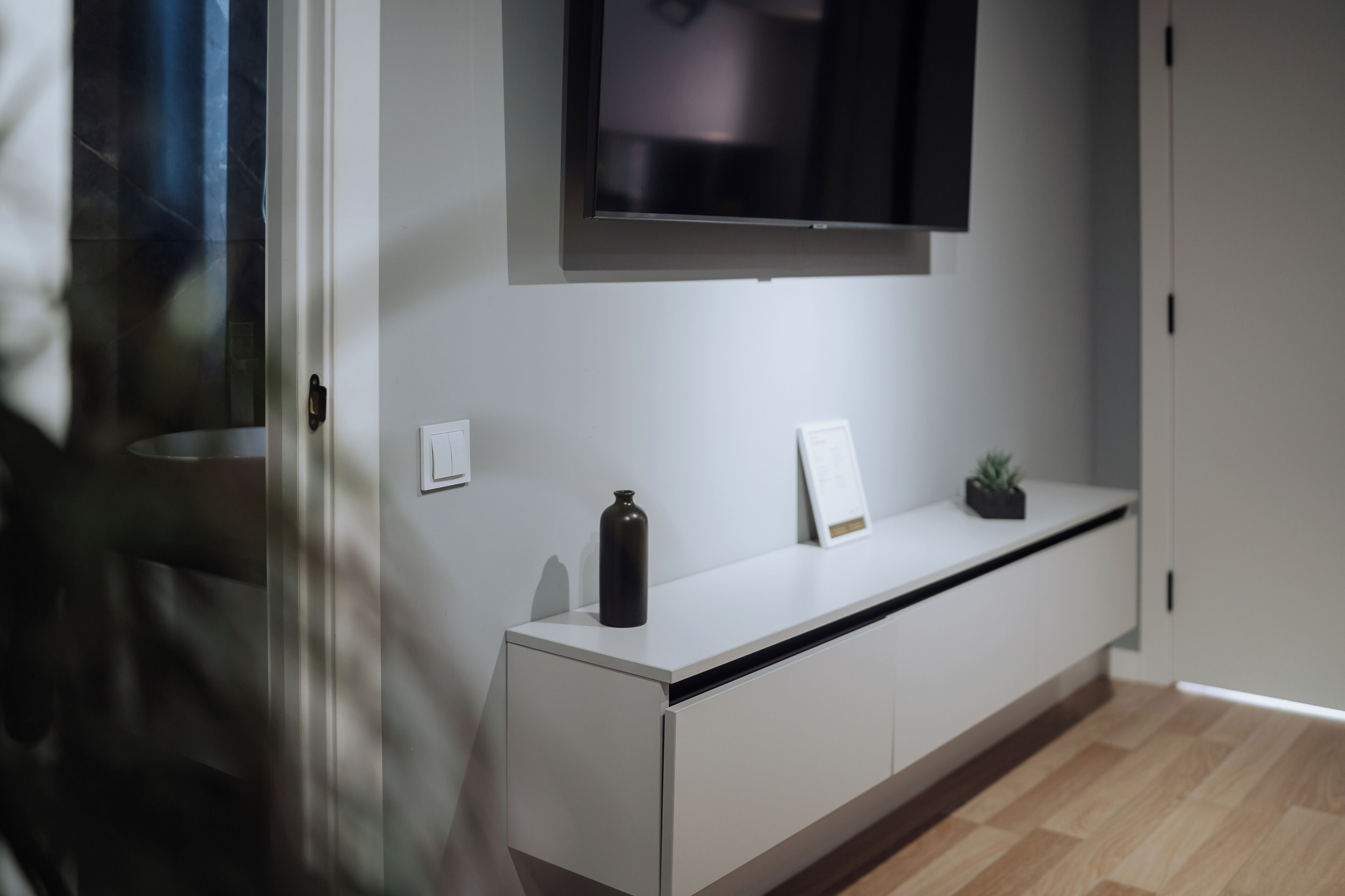 Quarto moderno e minimalista com uma mesa de consola branca, uma televisão de ecrã plano montada acima e uma pequena planta numa jardineira sobre a mesa.
