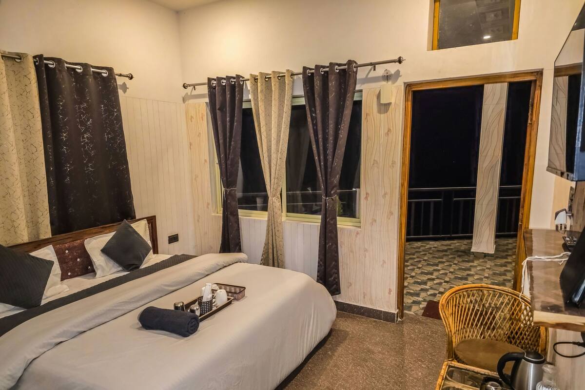 A hotel room with a bed, a small table with a kettle, a chair, and a balcony with a checkered floor.