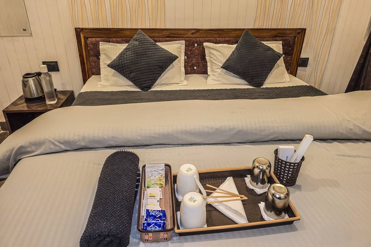 A hotel room with a bed, pillows, a tray with toiletries, and a nightstand with a kettle.