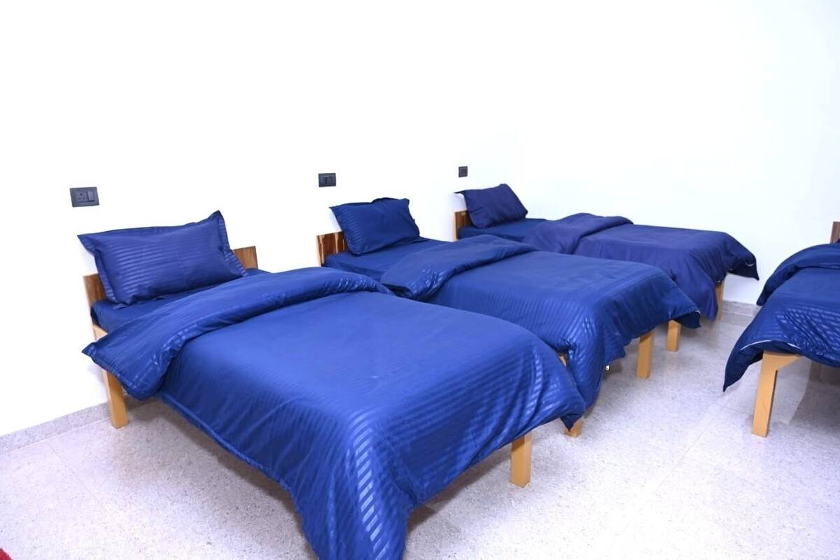 A row of four single beds with wooden frames and blue bedding, arranged against a white wall with electrical outlets.
