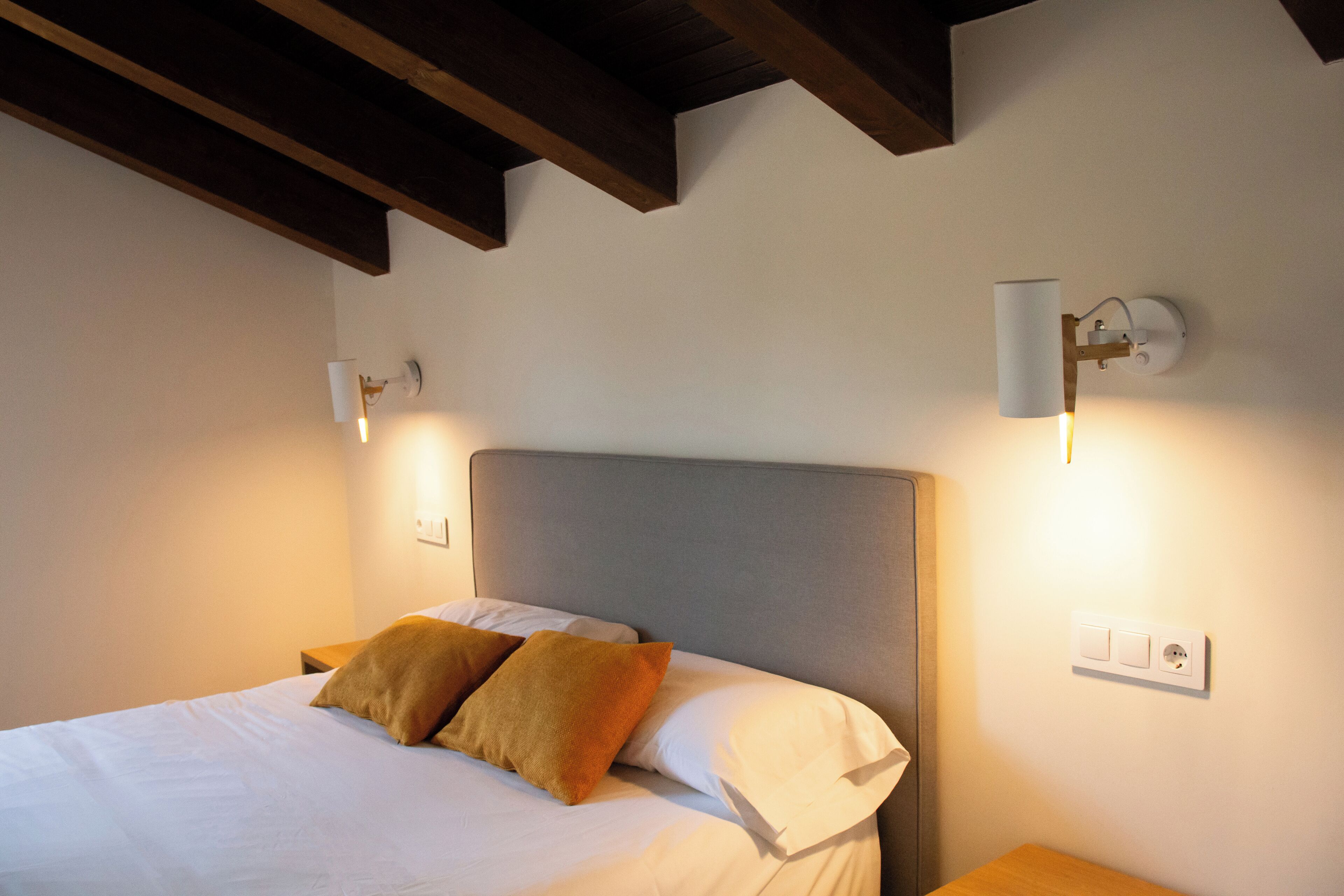 A neatly made bed with two pillows, a wooden nightstand, and a wall-mounted light fixture.