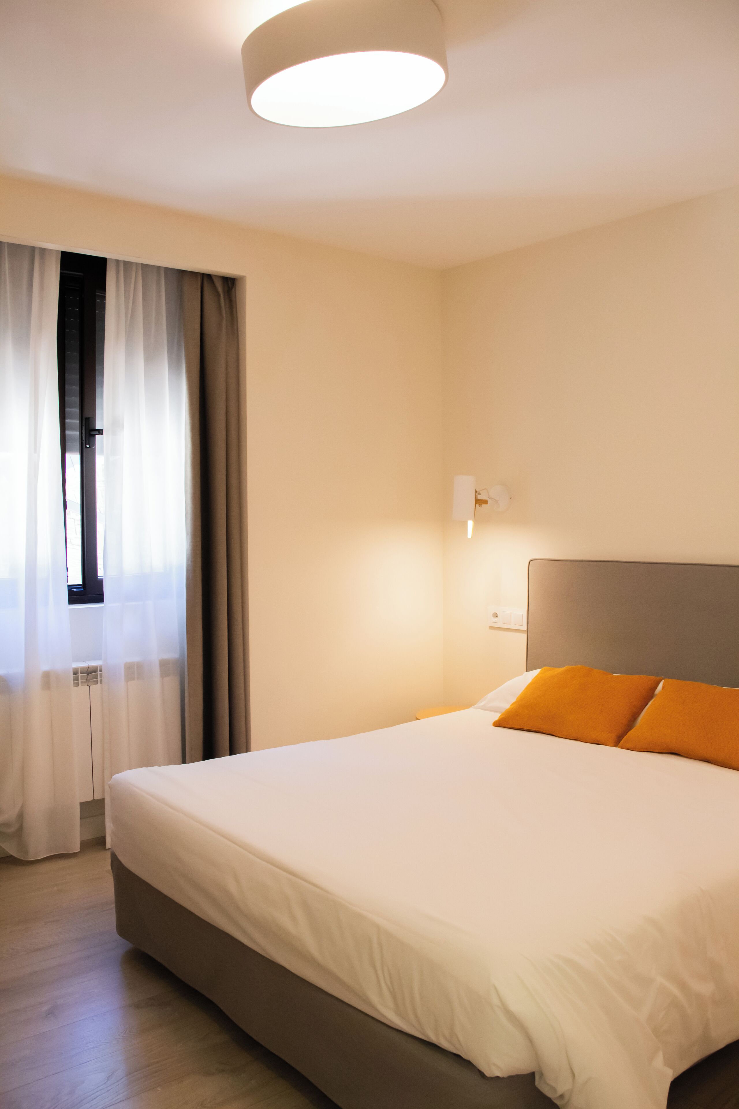 Quarto de hotel com cama grande, mesa de cabeceira, janela com cortinas e luminária fixa na parede.