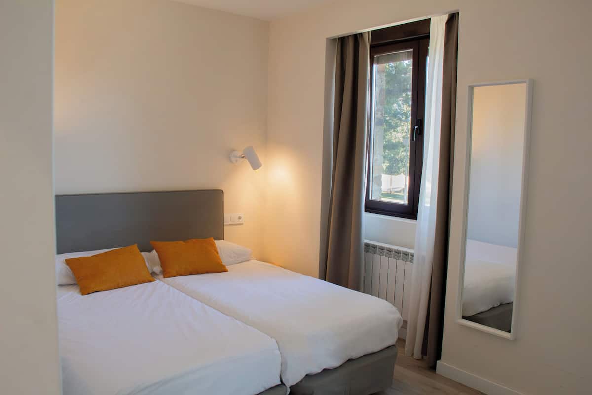 Ein Doppelbett mit weißer Bettwäsche und zwei orangenen Kissen, eine Nachttischlampe, ein Fenster mit Vorhängen und ein Spiegel an der Wand.