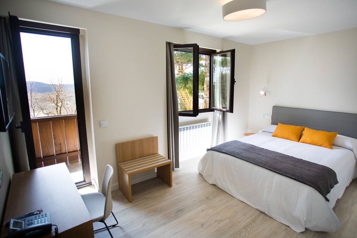 Ein Schlafzimmer mit Bett, Schreibtisch, Stuhl und einem Fenster mit Blick auf Bäume und ein Gebäude.
