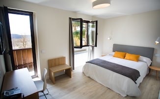 Un dormitorio con cama, escritorio, silla y ventana con vistas a árboles y un edificio.