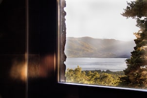 Vista a través de una ventana con un borde deshilachado, mostrando un paisaje con montañas, una masa de agua y árboles.