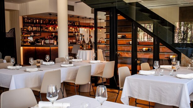 A restaurant with multiple tables set for dining, a bar area with shelves of bottles, and a staircase leading to an outdoor area. - Hotel Villalucerna (Vigo)