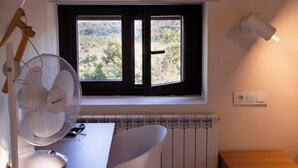 A room with a wooden desk, a white chair, a standing fan, a window with a view of greenery, and a radiator. - Hotel Villalucerna (Vigo)