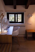 Una habitación con un escritorio de madera, una silla blanca, un ventilador de pie, una ventana con vistas a follaje y un radiador.