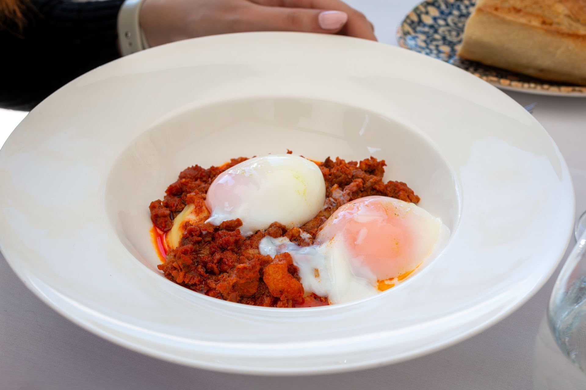 A white bowl with two poached eggs on a bed of red sauce, possibly a spicy meat dish, on a table.