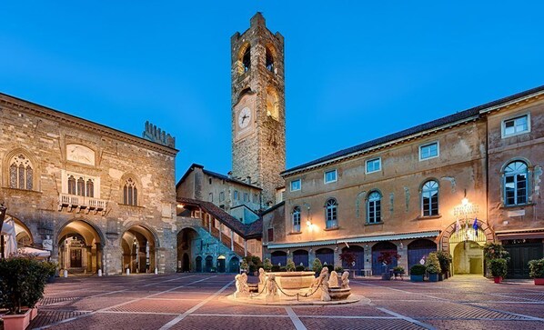 A historic square with a prominent clock tower, stone buildings, and a fountain. - LocandaMimmo (Bergamo)