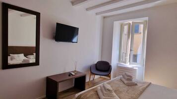 A hotel room with a bed, a TV mounted on the wall, a desk, a chair, and a window with blinds.