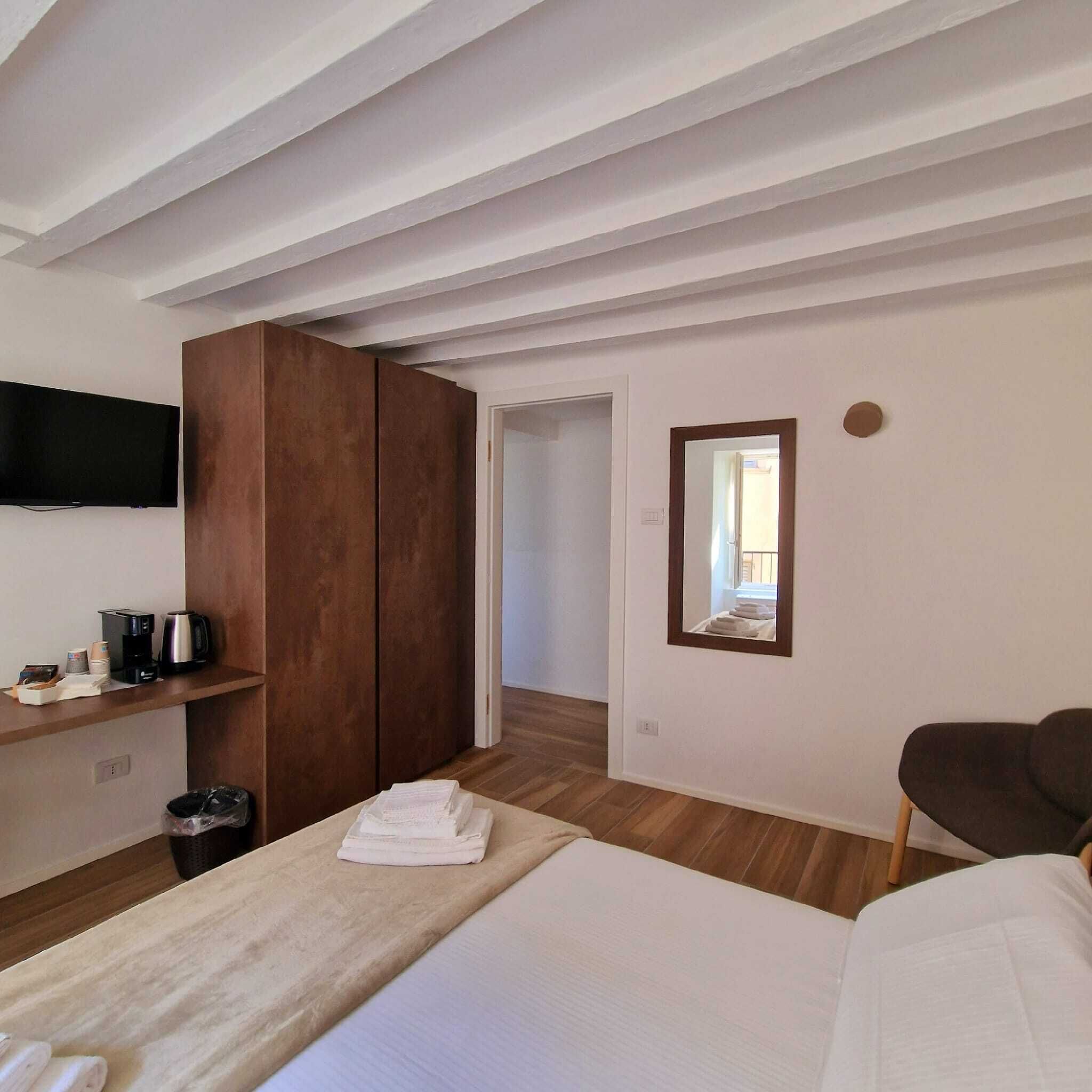 A hotel room with a bed, a wooden wardrobe, a television mounted on the wall, a small table with a coffee maker, and a mirror on the wall.