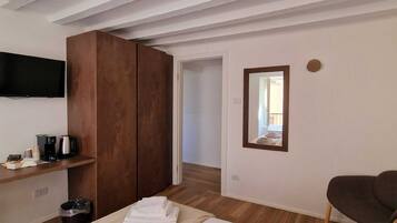 A hotel room with a bed, a wooden wardrobe, a television mounted on the wall, a small table with a coffee maker, and a mirror on the wall.