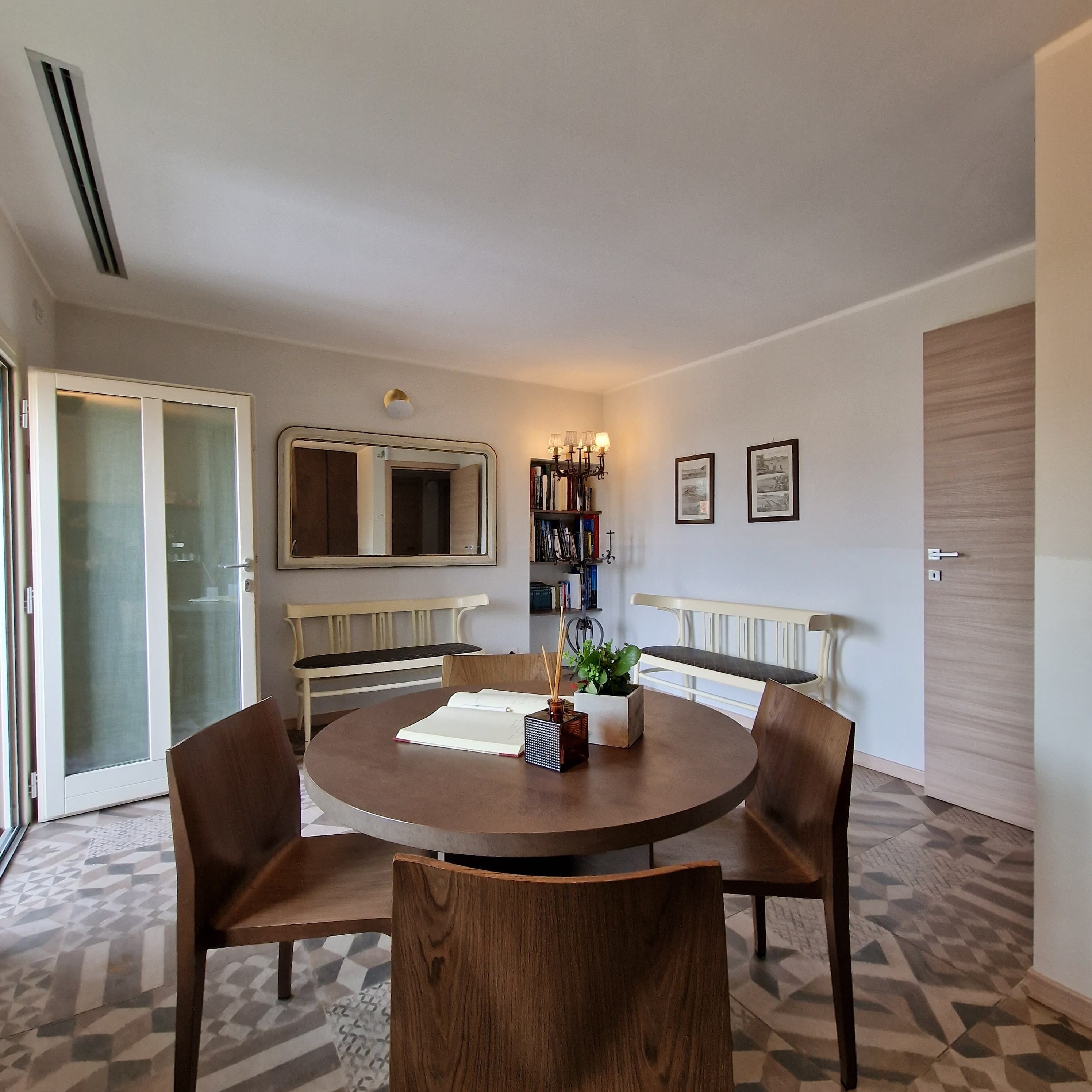 A dining area with a round table, four chairs, a bench, a bookshelf, and a door.