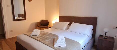 A neatly made bed with pillows and folded towels, a bedside table with a lamp, and a chair in a room with wooden flooring and a sloped ceiling.
