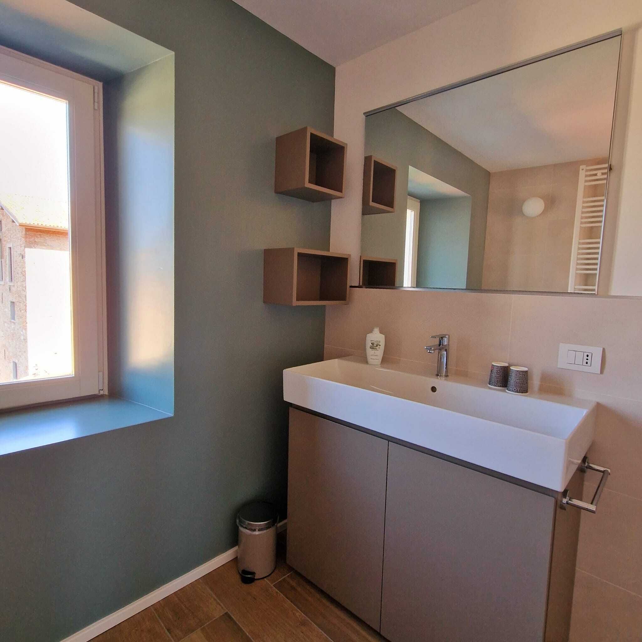 Ein Badezimmer mit einem großen Spiegel, einem weißen Waschbecken und einem Fenster. An der Wand befinden sich Holzregale und eine Mülleimer steht in der Ecke.