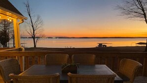A wooden deck with a table and chairs, overlooking a body of water during sunset.