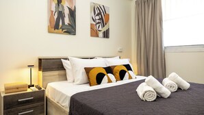 Un lit bien fait, recouvert d’une couvre-lit à motifs sombres, avec deux oreillers à dessins géométriques et une table de chevet sur laquelle se trouvent une lampe et un petit objet décoratif.
