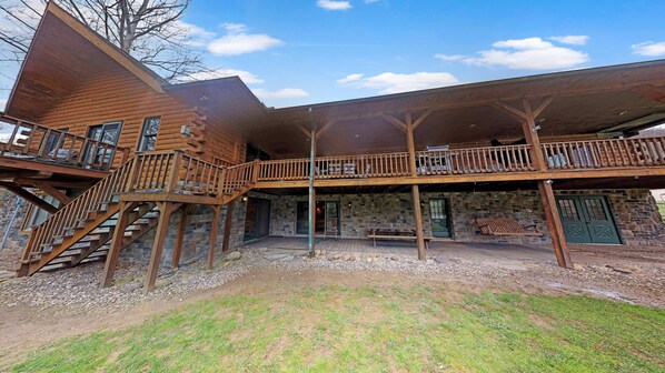A two-story log cabin with a stone foundation, a wooden deck, and a staircase leading to the upper level. - Sycamore Cabin with Hot Tub Breathtaking Views (Gaines)